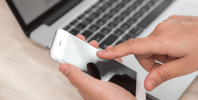 Pessoa usando smartphone em frente a um notebook, simbolizando o uso do MDM além do simples bloqueio de dispositivos.
