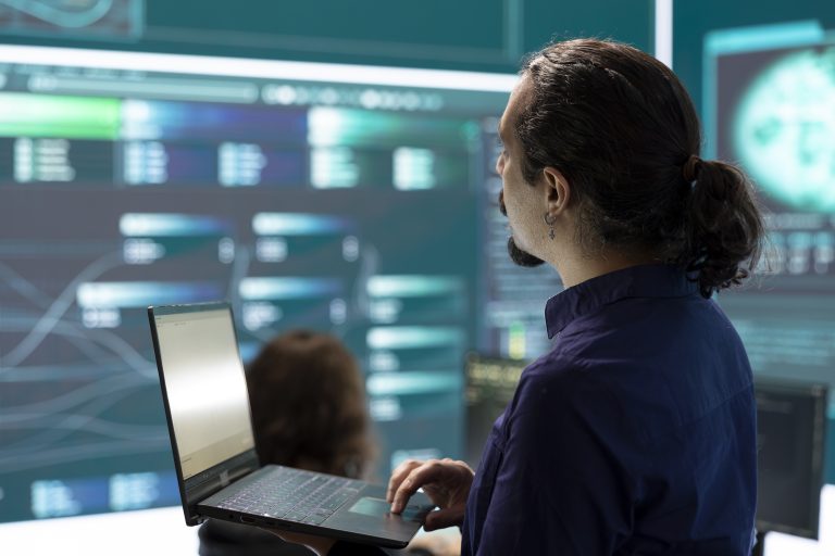 Profissional analisando informações em laptop diante de painéis digitais, representando monitoramento e segurança da informação em ambiente corporativo.