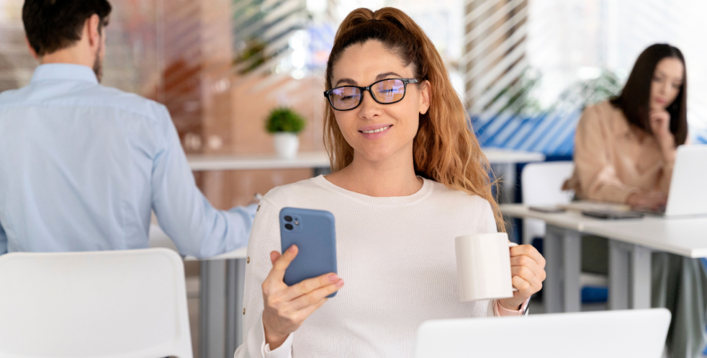 Mulher sorridente com óculos de grau e cabelos ondulados, sentada em uma mesa de escritório branca, segurando um smartphone azul em uma mão e uma caneca de café na outra. Há um laptop aberto e cadernos à sua frente.