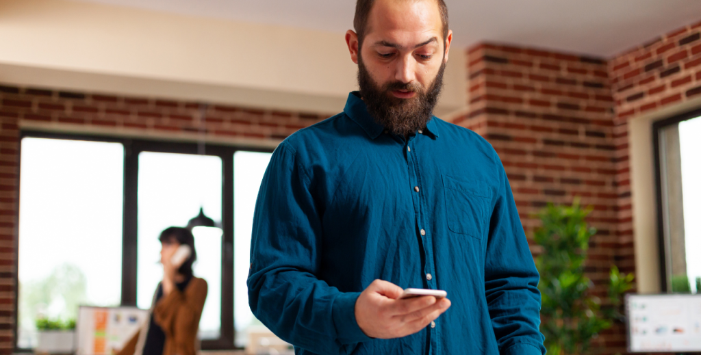 Homem barbudo com camisa azul em um escritório moderno, segurando um smartphone e olhando atentamente para a tela. Ao fundo, uma colega de trabalho fala ao telefone em um ambiente de startup com paredes de tijolos aparentes.